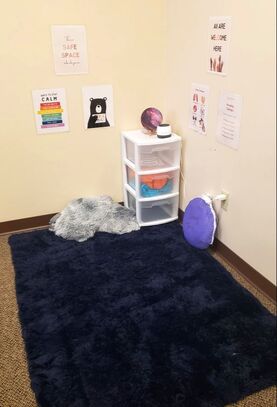 Picture of corner with shelf, posters on wall that say calm space, all our welcome here, blue carpet on floor, blanket, light and folded up tent.